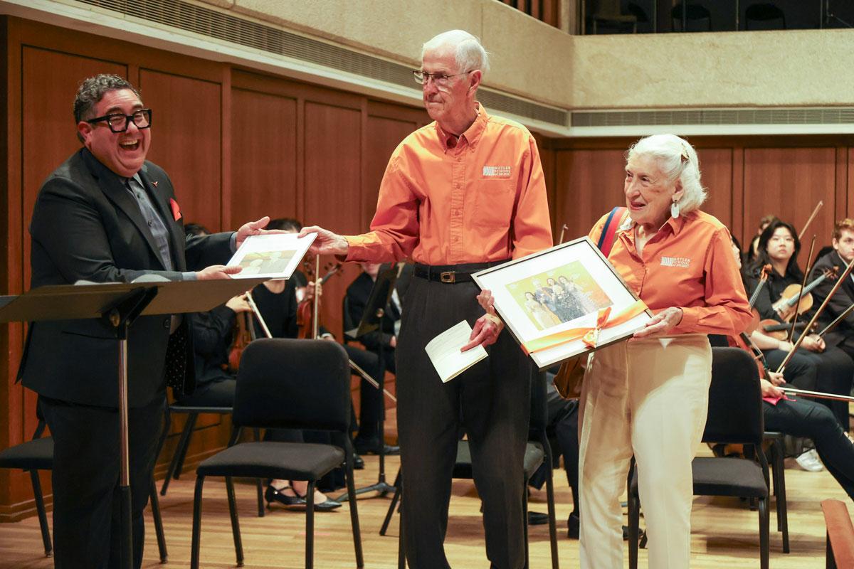 A man in a suit smiles as he hands a plaque to an older couple on a stage.