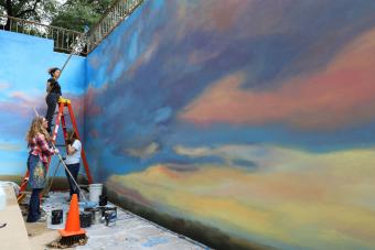 Three people paint a mural with sky and clouds on a concrete wall.
