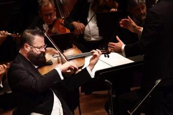 Violist Roger Myers in a tuxedo playing as a soloist in front of an orchestra, beside a conductor.