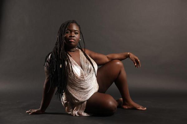 Daymé Arocena wears a slinky silver dress, sitting on the floor leaning on one hand.