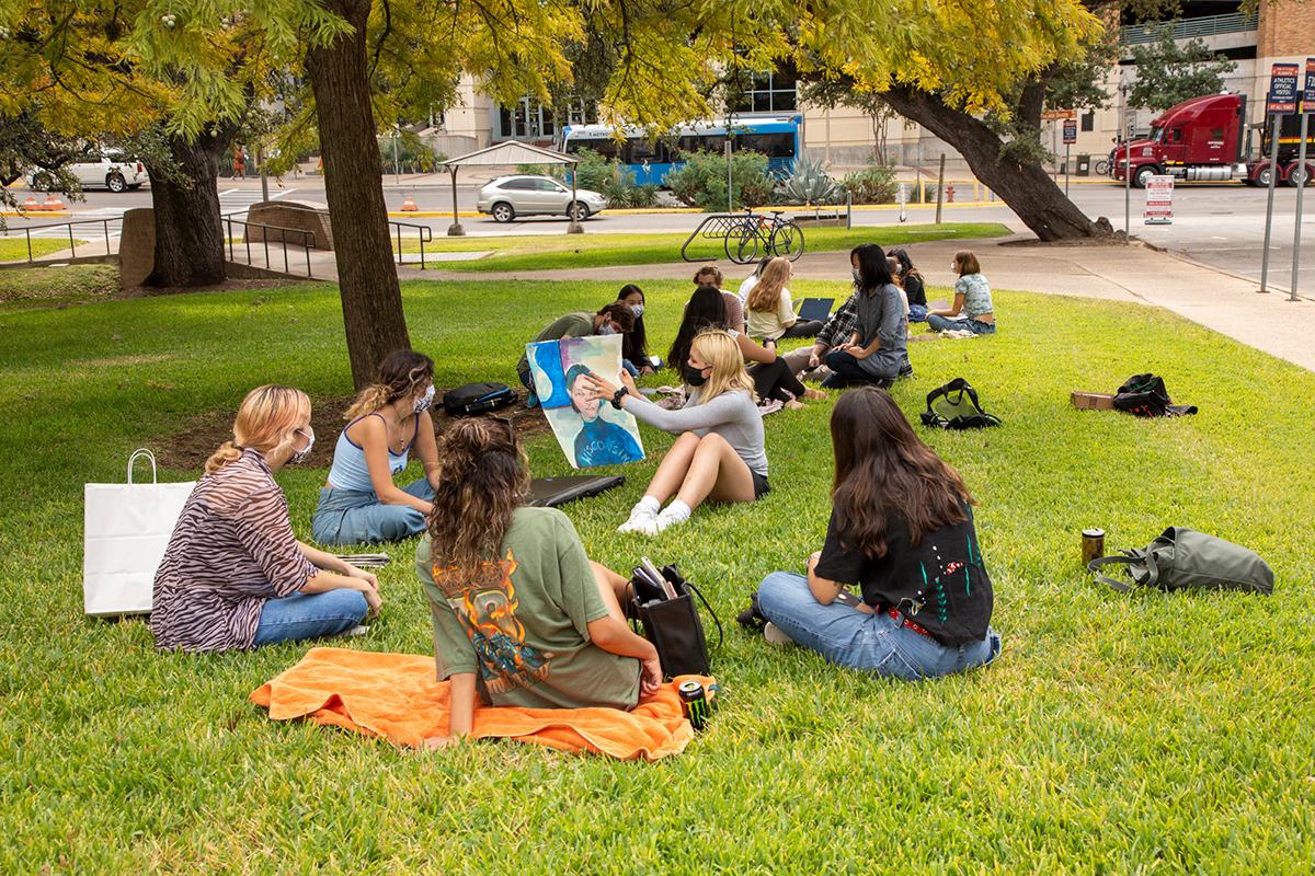 A group of masked students sit on a lawn at a safe social distance. 