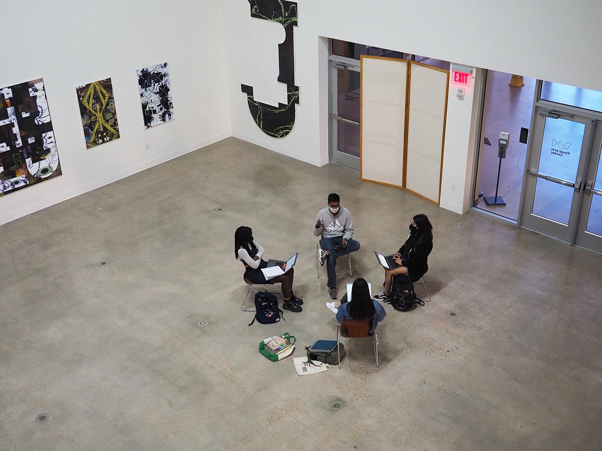 Masked students sit in a cluster of chairs in a large art gallery space. 