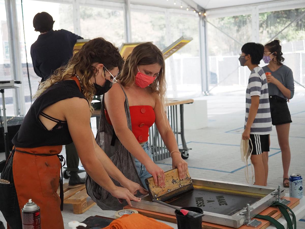 A masked student prints a t-shirt on a screenprinting press in an outdoor tent. 