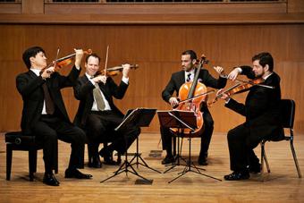 Four string players perform on a stage with wood paneling behind them.