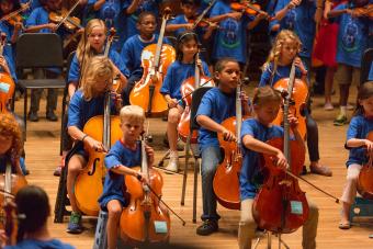 Young children in blue tee shirts play string instruments on a stage