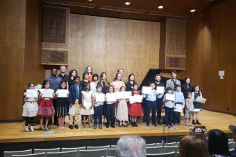 A group of Piano Project Students Stand on stage at a recital, proudly displaying their Piano Project Certificates
