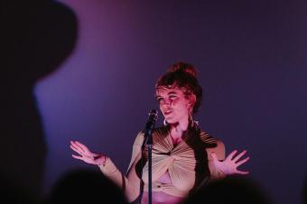 Katie Folger performs on stage behind a mic, holding her hands out while speaking