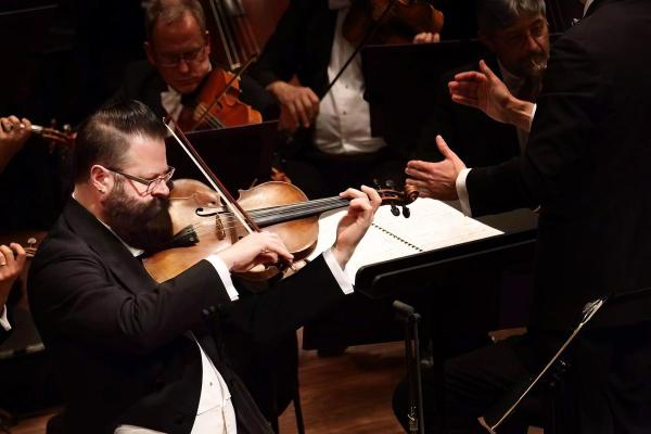 Violist Roger Myers in a tuxedo playing as a soloist in front of an orchestra, beside a conductor.