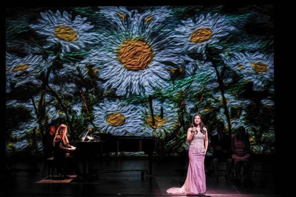 A singer in a pink gown and a pianist perform in front of a backdrop of daisies.