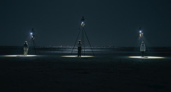 film still of scene on beach at night with individuals perched atop three large steel tripods shinin