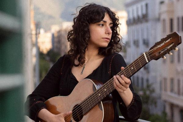 Singer/songwriter Amanda Pascali holds a guitar in front of a backdrop of buildings.