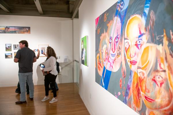 A painting of faces on a gallery wall with visitors in the space