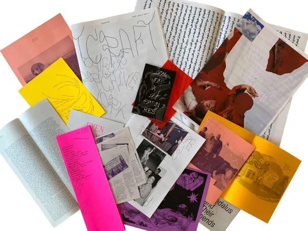 An assortment of zines and printed materials spread out in an overlapping arrangement. The collection includes various colors such as bright pink, yellow, orange, and red, along with black-and-white pages. Some zines feature handwritten elements, abstract illustrations, and photographs, including an image of a person wearing red and another of a group of people. The text on the covers and pages suggests themes of queer archival work and artistic expression.