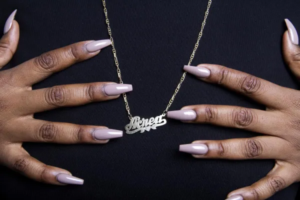 A close-up photograph of a person’s hands with long, glossy, nude-colored nails gently holding a gold nameplate necklace that reads 'Acead' in script font. The necklace has a small heart detail and hangs against a black fabric background.