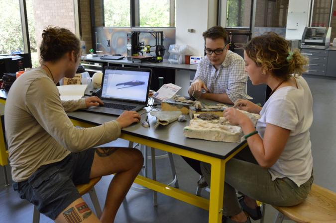 Three students work at a table together in the UT 3D printing space.