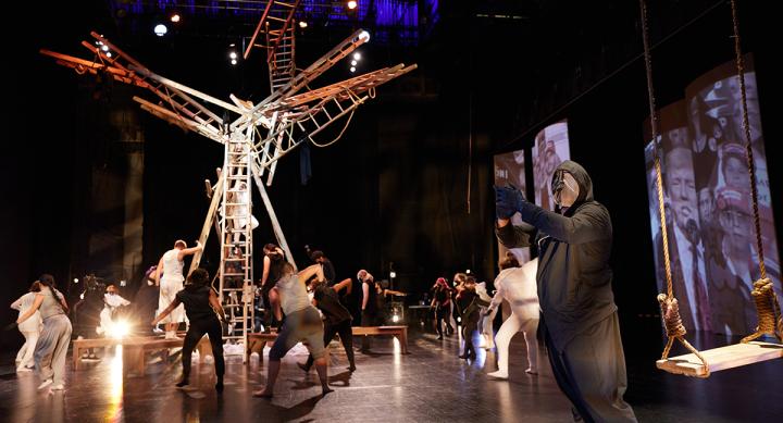 A stage with a structure comprised off ladders, with dancers in black and white hoods and masks.