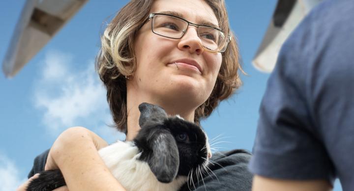 A young student smiles in front of a blue sky while holding a rabbit.