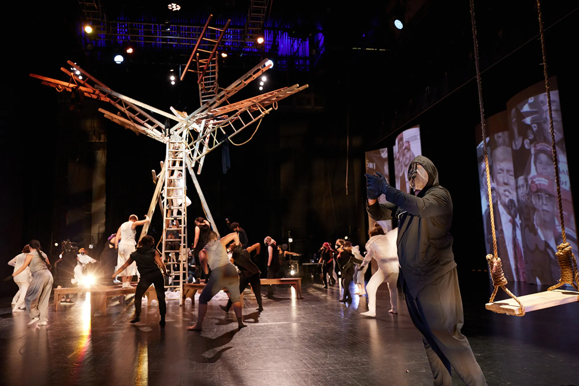 A stage with a structure comprised off ladders, with dancers in black and white hoods and masks.