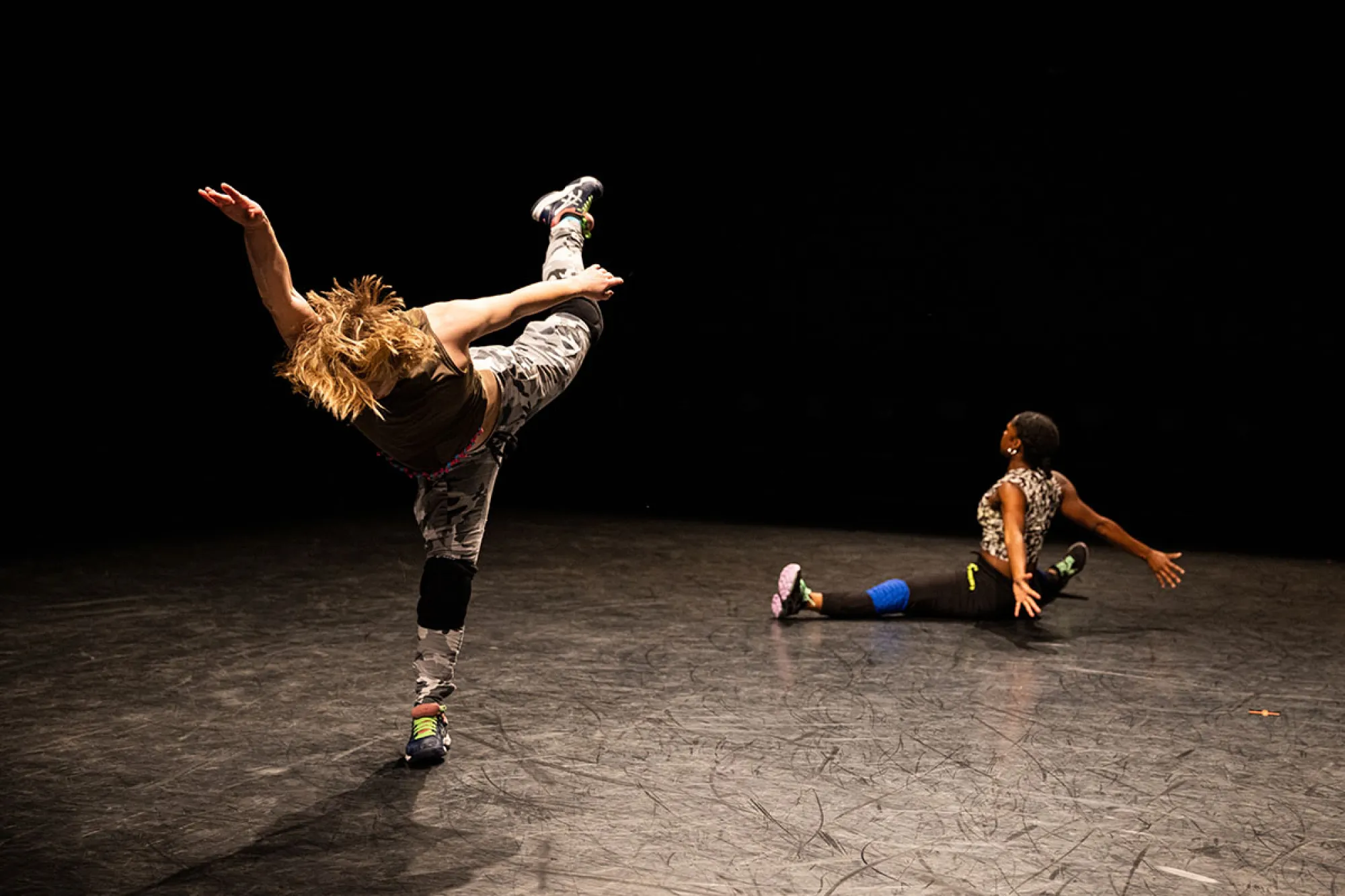 A dancer in street clothes balances one leg, while another is seated with her legs spread wide.