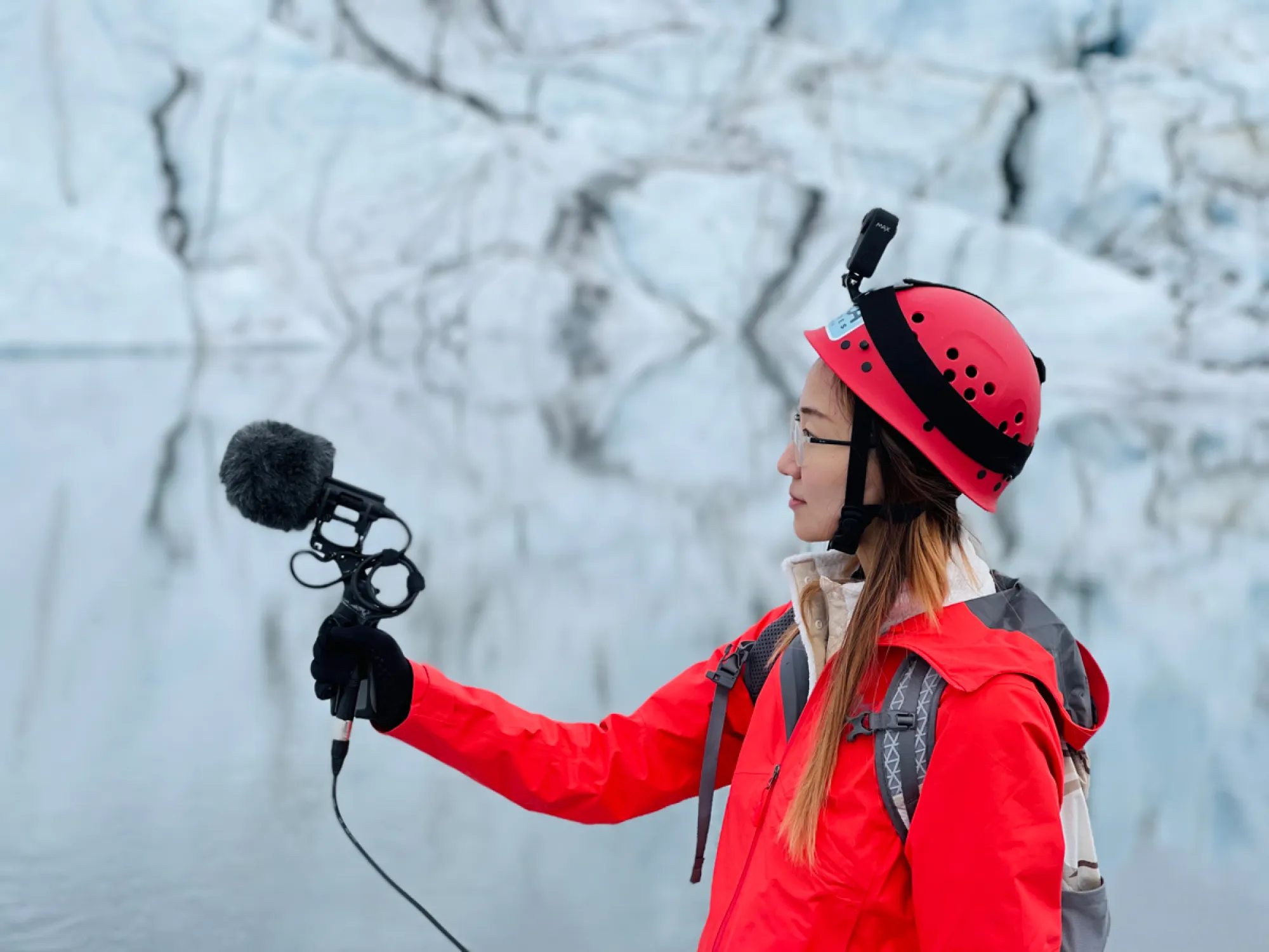 A woman in a heavy coat holds a microphone up in front of a glacier.