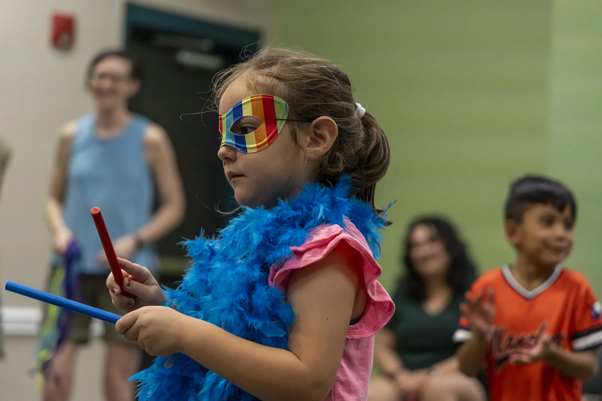 A child wearing a mask and blue feather boa plays percussion sticks at a Meet the Opera program