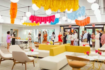 A group of mostly women, wearing masks, gathers inside a space decorated with bright colors and natural light