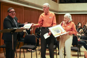 A man in a suit smiles as he hands a plaque to an older couple on a stage.