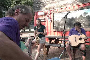 A person wearing a skirt plays a saxophone, flanked by a person playing an acoustic guitar and a bass. 