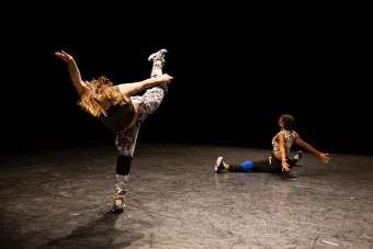 A dancer in street clothes balances one leg, while another is seated with her legs spread wide.