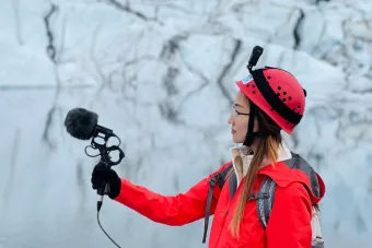 A woman in a heavy coat holds a microphone up in front of a glacier.