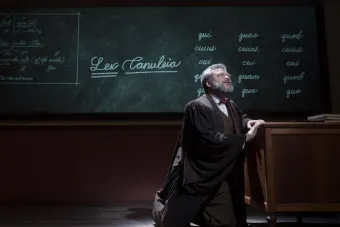 A still from the film Goodbye, Mr. Chips, showing a man kneeling beside a desk in front of a chalkboard.