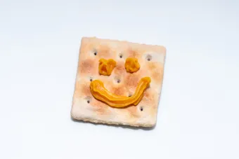 A saltine with a smiley face made from Cheez Whiz against a neutral background