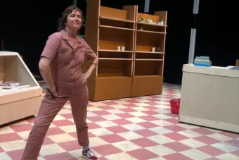 Photo of Lisa Damour standing in a pale red boiler suit on a checkered linoleum floor part of the grocery store set of her play frozen section