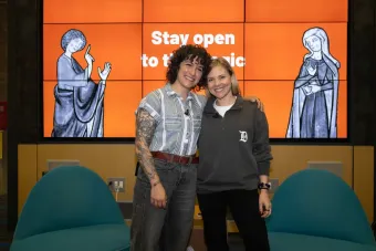 Two smiling speakers stand arm-in-arm in front of a large digital display with the words “Stay open to the magic” and medieval-style illustrations of gesturing figures.