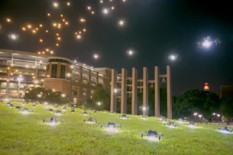 Dozens of drones line a grassy hillside on the University of Texas at Austin campus at night, with more drones ascending into the sky above university buildings and the Tower glowing in the distance.
