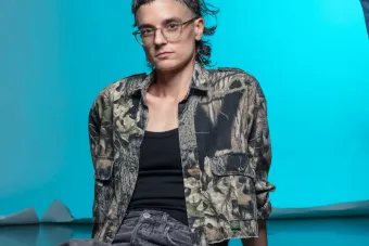 riel Sturchio sitting on the floor in a studio with a blue background. Their hair is being fixed by a disembodied hand (the hand of friend Dr. Kanwal Matharu). The photo was taken by Thalassa Raasch.
