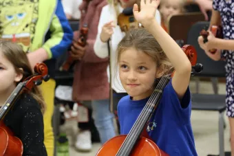 A child holds a cello in class and is raising their hand