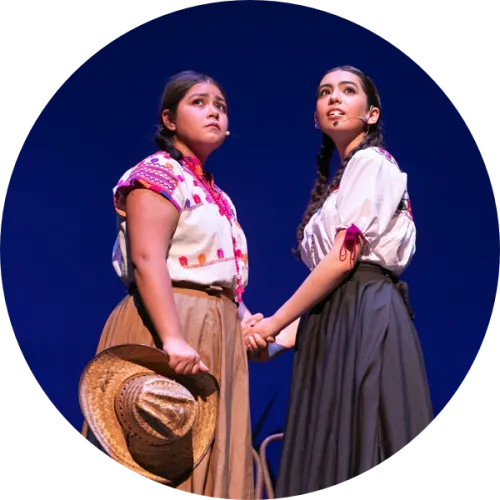 Two actresses in period costume clasp hands on stage.
