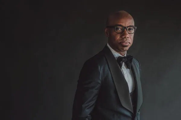 A portrait of choral conductor Jason Max Ferdinand wearing a black tuxedo, glasses and a bowtie against a black background.