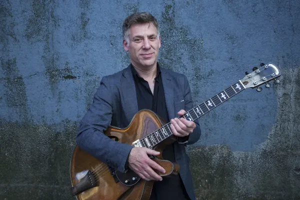 Jazz guitarist Peter Bernstein stands in front of a blue painted cement wall holding his guitar