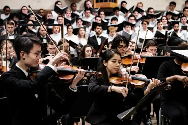 The first violin section of the University of Texas Symphony Orchestra plays, foregrounded by University of Texas choral singers.