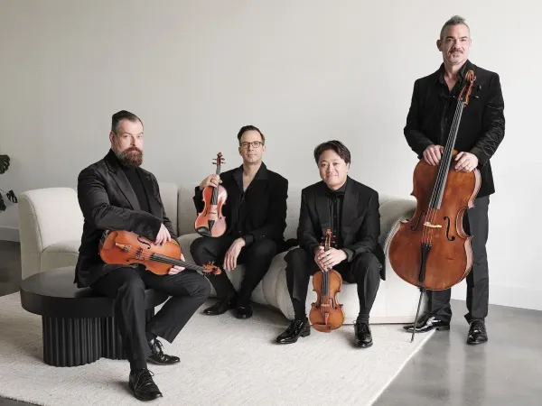 Members of the Miró Quartet wear black suits and gather around a white couch holding their instruments.