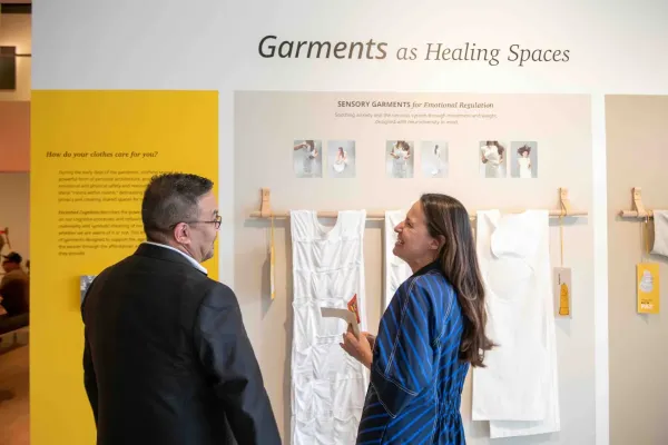A woman in a blue striped dress speaks with a man in front of an exhibition display titled "Garments as Healing Spaces." The exhibit features white sensory garments designed for emotional regulation and panels explaining the concept of clothing as personal architecture for mental and emotional care.