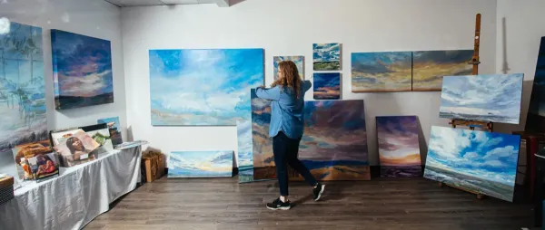 Artist arranging large landscape paintings in a studio gallery space, surrounded by vivid skies and cloudscapes displayed on easels and walls.