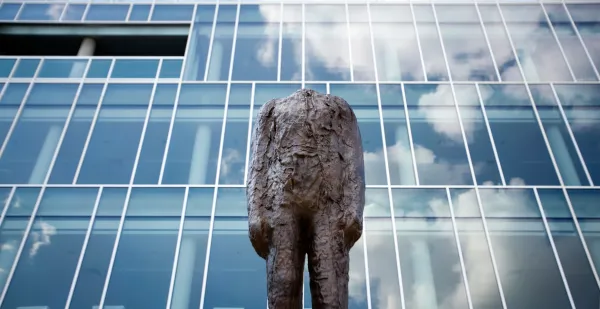 A headless bronze statue behind a building made of panes of glass which reflect the sky.