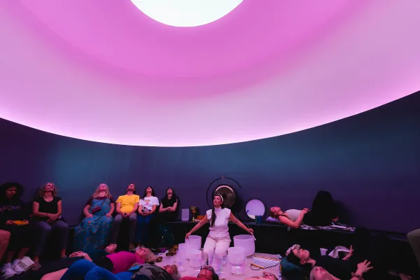 A woman sits in a curved room with purple lighting and an oculus opening to a white sky above her. While she singing bowls of various sizes, she is surrounded by a group of about 15 people who are all looking up.