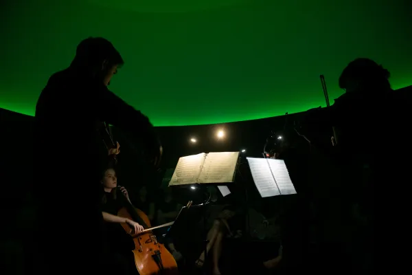The silhouette of two violinists performing in a dark curved room lit by a green light.