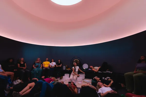 A women plays singing bowls while surrounded by sitting and laying listeners. She sits in a curved room lit with pink light underneath an oculus in the ceiling.
