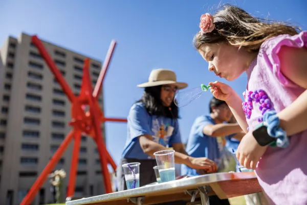 A young girl blows a soap bubble outside in front of a large red metal sculpture.