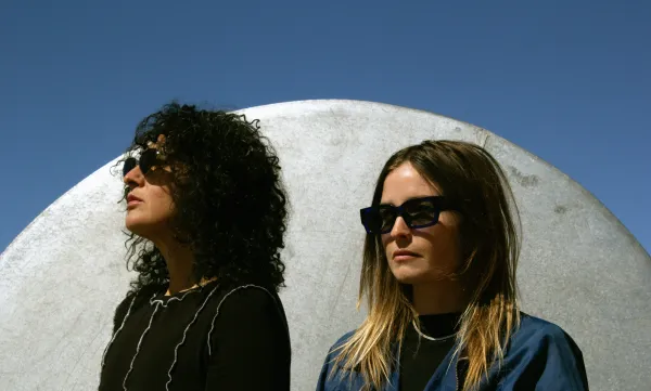 A portrait of two women standing side by side wearing sunglasses. They are outside, and behind them is a cement dome.